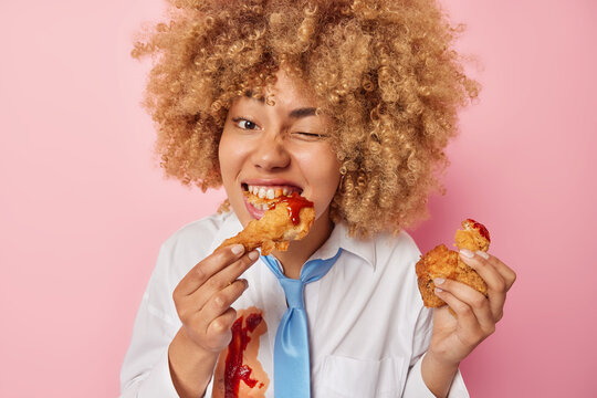 Photo of hungry young European woman eats delicious fried chicken nuggets with ketchup winks eye enjoys eating fatty appetizing unhealthy food dressed in formal clothes isolated over pink background