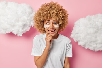 Photo of surprised curly haired woman bites finger applies beauty patches under eyes for reducing bags wears casual white t shirt poses against pink background clouds around. Skin care procedures