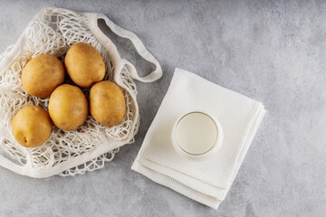Potato milk and raw potatoes in a mesh bag. Alternative non dairy drink and ingredients on gray background. Allergens free and zero waste concept. Top view. Copy space