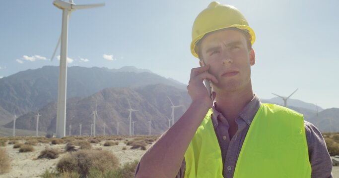 Close up on technician in hard hat and wearing yellow high-visibility vest, at wind farm, listening on a phone call. Wind turbines and mountains in background
