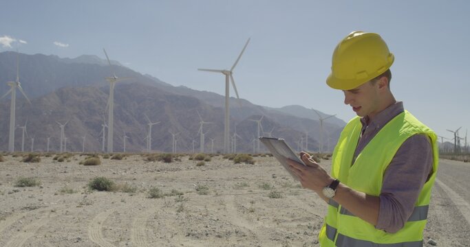 Wide view, technician in hard hat and wearing yellow high-visibility vest, looks up to camera while using a tablet computer and stylus at a wind farm. Road leads towards wind turbines and mountains in