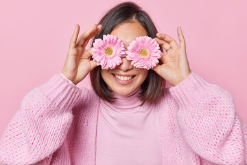 Obraz na płótnie Canvas Horizontal shot of pleased unrecognizable dark haired woman covers eyes with gerbera flowers smiles toothily enjoys pleasant fragrant dressed in casual knitted jumper isolated over pink background