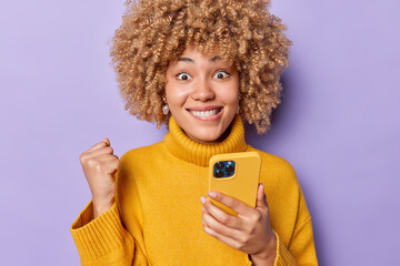 Cheerful beautiful woman with curly hair celebrates victory clenches fist expresses success smiles toothily wears yellow warm jumper isolated over purple background views content in cellular screen