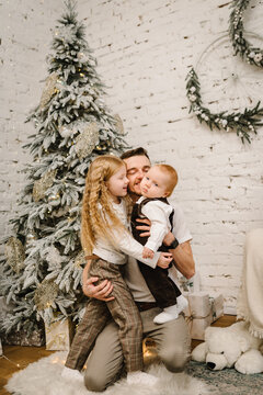 Father hugs son and daughter near Christmas tree. New Year's holidays. Dad embraces kids in festively decorated room and decorations. Decorated interior of house. Concept of family holiday. Closeup.