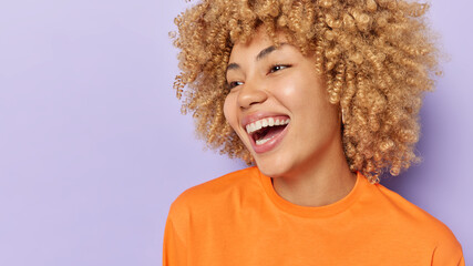 Horizontal shot of pretty cheerful woman with curly hair smiles broadly shows white teeth wears...