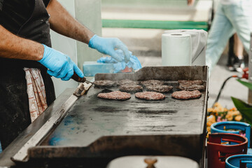 A man fries burger meatballs. Man grills some kind of marinated meat on gas grill during summer time. Meat on the grill is cooked by man's hand in a blue glove.