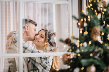 Christmas tree with garlands. Man kissing woman in decorated interior room. Couple enjoying winter...