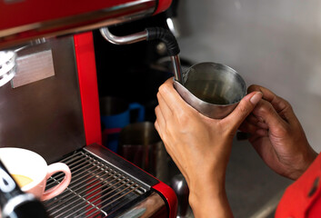 hands of a barista holding the metal jug where she is heating the milk for her customer's coffee. the machine and her uniform are red. there is a colorful cup with coffee. vertical photograph.