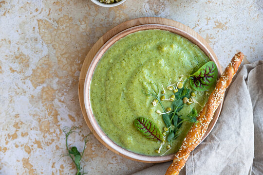 Green vegetable cream soup served with micro greens, salad leaves and breadstick on concrete background. Vegan soup puree of green vegetables. Diet food concept. Top view.