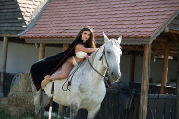 sensual brunette princess with sexy white bikinis riding a horse . Portrait of a girl  with cape...