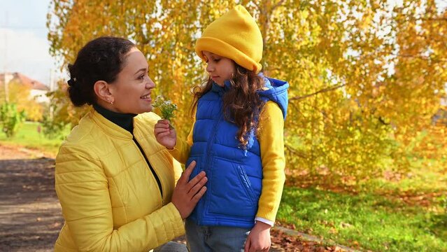 Dark-haired multi-ethnic pretty woman, a loving mother and her daughter in bright blue and yellow jackets, enjoying a weekend together in a beautiful autumn park on a sunny warm day