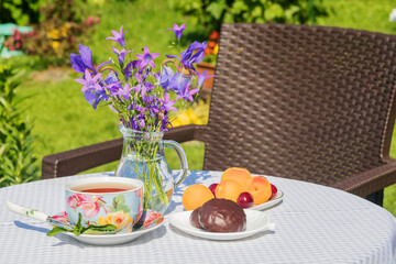 On the garden table, a cup of tea, chocolate marshmallows, fresh fruits. A bouquet of flowers in a vase.