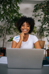 Vertical portrait of laughing curly black young woman holding pen in hand, smiling looking at camera sitting at table in light home office room with modern biophilic interior design.