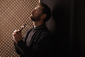 Catholic priest in cassock holding cross and praying to God in confessional, space for text - obrazy, fototapety, plakaty