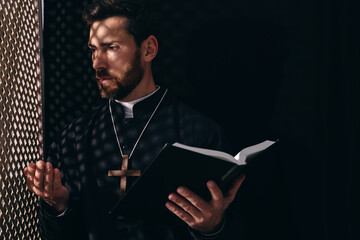 Catholic priest in cassock holding Bible and talking to parishioner in confessional - obrazy, fototapety, plakaty