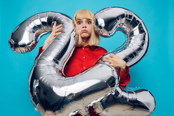 a happy, enthusiastic woman in a red shirt stands on a blue background and holds inflatable balloons in the shape of the number twenty-two in silver color, amusingly inflating her cheeks