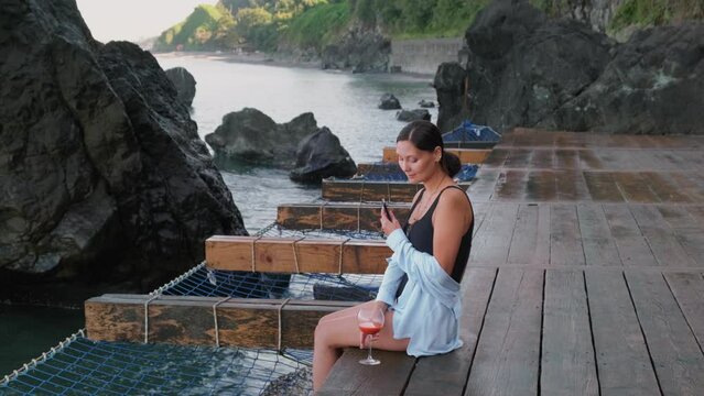 Woman Using Smartphone and Relaxing at the Sea. Female Wrights Messages on the Phone and Enjoying with Glass of Juice on the Rocky Beach.