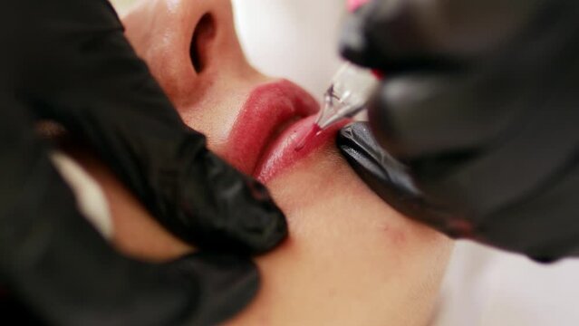 Permanent makeup procedure applying rose red pigment using tattoo machine in beautician salon. Microblading tattoo lips, close up	