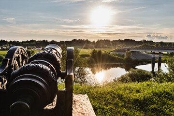 An outdated cast-iron cannon on the river bank is directed towards the settlement. European historic site during sunset in summer