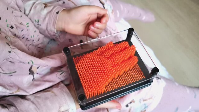 Young girl playing with plastic pin art toy. Orange pins in a black frame and hand inprint touch relaxing game. Child in pink dress hold antistress 3D pinscreen game video