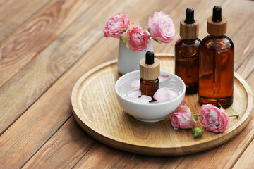 Bottles of rose essential oil and flowers on wooden table, space for text