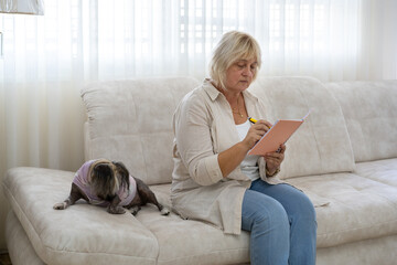Beautiful elderly woman sitting on the couch in living room and writing in planner.