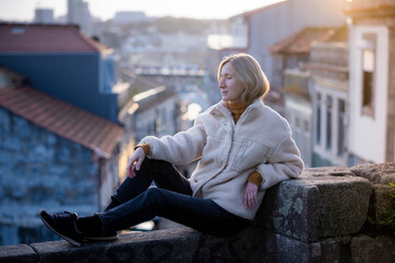 Fototapeta na wymiar A female tourist sits on a masonry wall in an old European city. The houses in the background are blurred.