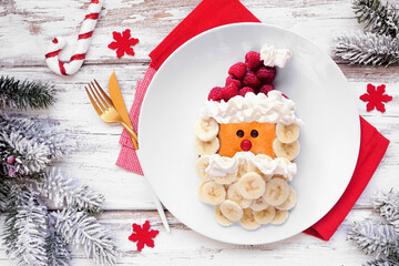 Fun Christmas Santa Claus pancake on a white plate with raspberry hat and banana beard. Overhead...