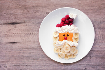 Cute Christmas Santa Claus pancake on a white plate with raspberry hat and banana beard. Overhead...