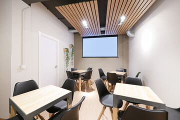 Dining room of a restaurant with square beech wood and gray metal tables and a projection screen on...