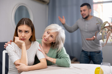 Offended woman and her senior mother sitting at table at home, her husband standing behind and swearing at her.