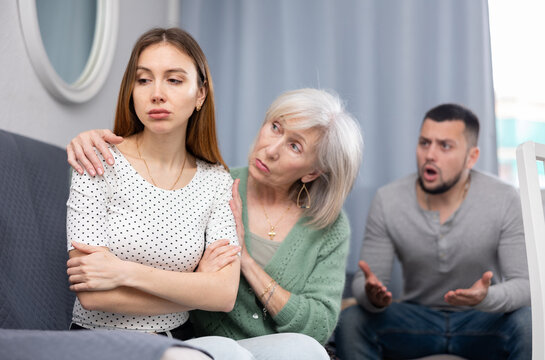 Portrait of sad young adult woman sitting at home while her husband berating her. Elderly woman her mother embracing and soothing