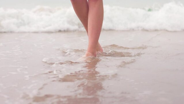 Close-up shot of female feet walking along sand, washed with foamy waves. Romantic summer vacation destination concept as seen from the shore. High quality 4k footage