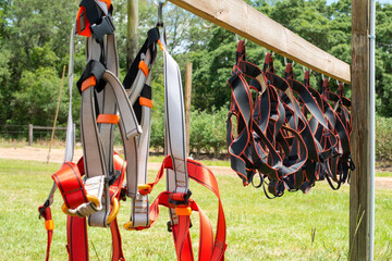 A wooden rack with multiple white, black and orange colored rock climbing equipment, cords, harnesses, hooks, fasteners, cams, belts, slings and cables hanging from a board outside in a grassy field. 