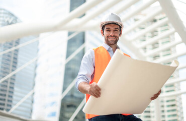 Fototapeta premium Young caucasian man holding a big paper, a guy wearing a light blue shirt and jeans with an orange vest and white helmet for security in the construction area.