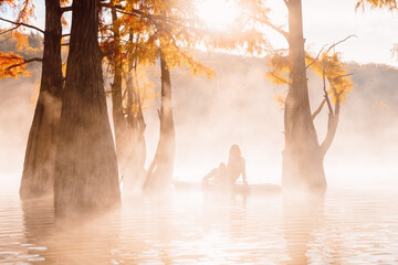 Woman relaxing on paddle board at the lake with morning fog, sun and autumnal Taxodium trees