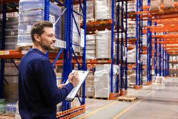 Happy manager holding clipboard in warehouse with lots of products. Space for text