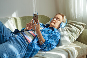 Mature woman in headphones using smartphone while lying on sofa at home