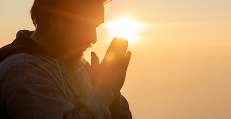Close up portrait of young adult male with beard with praying hands praying for thank god golden sunset background. Concept of religion faith - obrazy, fototapety, plakaty