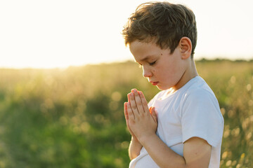 Boy closed her eyes and praying in a field at sunset. Hands folded in prayer concept for faith,...