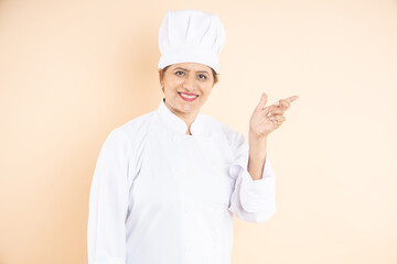 Happy mature female chef or cook dressed in uniform point finger at blank space isolated on beige...