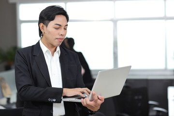 Attractive asian man employee worker in formal dress standing, using computer and looking at camera with coworker at workplace. Businessman smiling and working with notebook in the office.