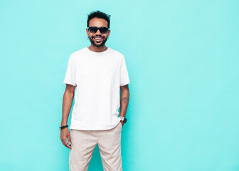 Handsome hipster smiling model. Sexy unshaven man dressed in white summer t-shirt and jeans clothes. Fashion male posing near blue wall in studio. Isolated. In sunglasses