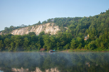 chalk mountain overgrown with dense vegetation, at the foot of the river in the morning haze and a...