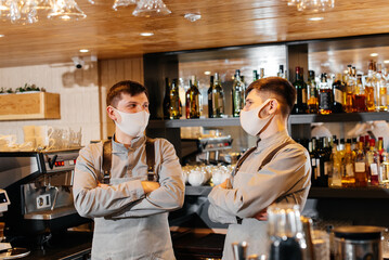 Two stylish bartenders in masks and uniforms during the pandemic, stand behind the bar. The work of restaurants and cafes during the pandemic.
