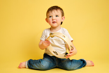 Toddler baby plays the tambourine, a child with a percussion musical instrument on a studio yellow background. Happy child musician playing hand drum. Kid aged one year four months