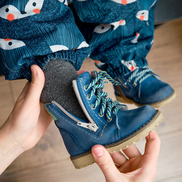 The Mother Puts A Blue Shoes On The Foot Toddler Baby Sitting In The Home Hallway. Woman Mom Dressing Warm Boots Clothes On Child For Winter Walk In Cold Weather. Kid Aged One Year And Three Months