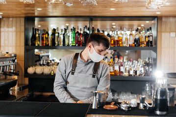 A stylish bartender in a mask and uniform during a pandemic is preparing cocktails at a party. The work of restaurants and cafes during the pandemic.