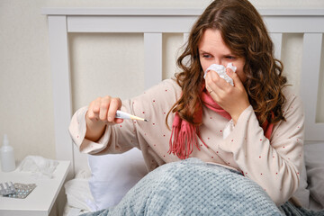 A sick woman blows her nose into a napkin with a thermometer in her hand, fever and high...