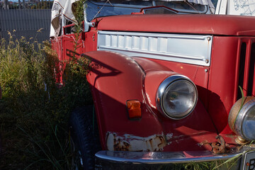 Part of an old car. An old red car is parked in the grass. The car is covered with a white cloth from the sun. You can see chrome parts, headlight, bumper, hood, horn. Selective focus.